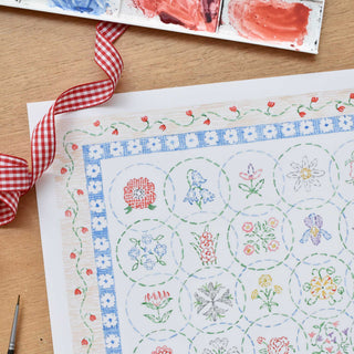 Detail of watercolor embroidery pattern with floral designs on fine paper, styled with red checkered ribbon, and paintbrush on wooden surface