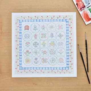 Cross-stitch embroidery print with floral designs on a white paper, placed on a wooden surface with watercolor paints and brushes, painted and styled by Sally King McBride of The Letter Nest