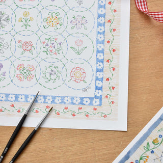 Detail of artist's signature on a watercolor Flower Embroidery pattern styled on a wooden surface with gingham ribbon and paintbrushes
