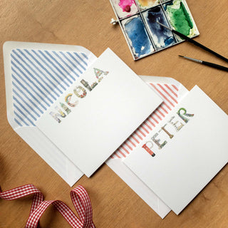 London Personalized Stationery by The Letter Nest in names "Nicola" and "Peter" spelled in watercolor-printed London letters with blue and red envelope liners styled in a flatlay with paints and ribbon