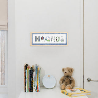 A nursery art print featuring the name "Magnus" in Sports-themed watercolor letters with a blue gingham border and natural wood frame hangs on a white wall above a white shelf with books, a white plate, and a teddy bear in a decorated nursery